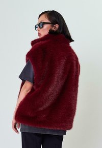 Burgundi spalvos dirbtinio kailio liemenė su aukštu apykakle, turinti tekstūruotą paviršių. Dėvima ant tamsios marškinės ir derinama su juodais stačiakampiais akiniais nuo saulės.