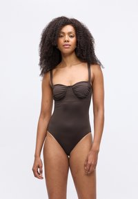 Donna con capelli ricci che indossa un costume da bagno attillato, intero di colore marrone scuro con spalline sottili, su uno sfondo bianco semplice.