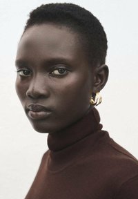 Pulover maro cu guler înalt, portret apropiat cu păr scurt și cercei aurii rotunzi pe fundal deschis. Textură netedă a pielii.