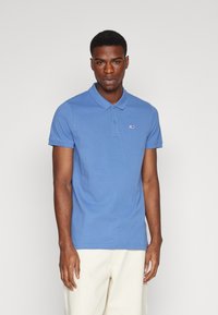 Camisa polo de algodón azul con mangas cortas y cuello. Presenta un pequeño logo en el pecho. Se lleva con pantalones cortos de color claro.