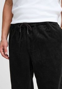 Pantalones de pana negra con acanalado vertical, que cuentan con una cintura elástica y cierre de cordón. Incluye puntas metálicas en el cordón.