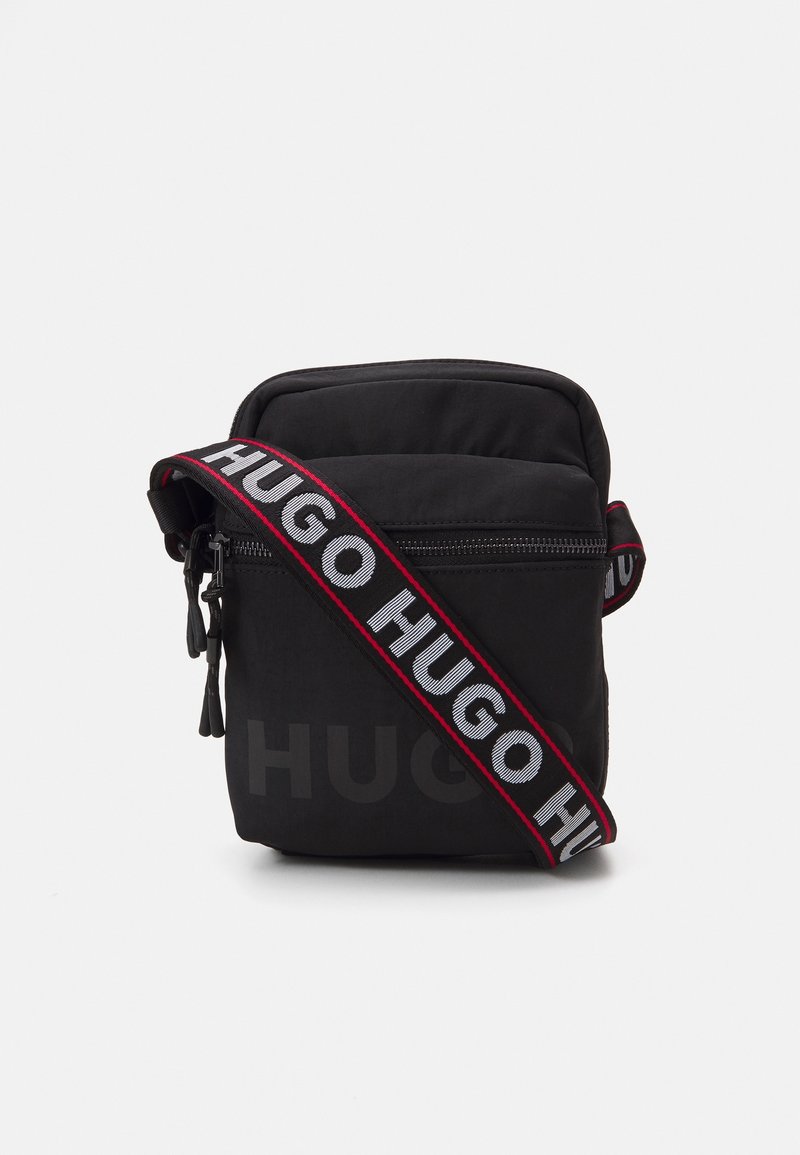 Musta nailoninen olkalaukku pehmustetulla muotoilulla, jossa on vahva säädettävä hihna, jossa on "HUGO"-logo valkoisella ja punaisella. Vetoketjukiinnitys.