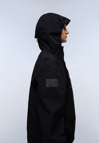 Persona che indossa una giacca nera con cappuccio e un sottile emblema di bandiera sulla manica superiore, rivolta a destra su uno sfondo chiaro e uniforme.