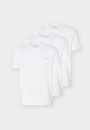 Άνδρας με αφροχτένισμα, φοράει λευκό μπλουζάκι, ριγέ πουλόβερ περασμένο στους ώμους, μπλε τζιν και καφέ σανδάλια, στέκεται με το ένα χέρι στην τσέπη.