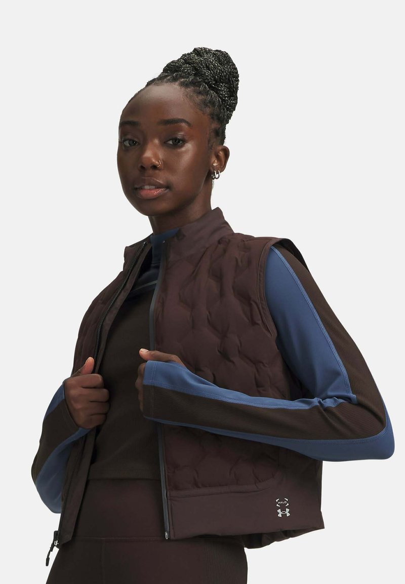 Woman with braided hair wears a brown quilted vest over a blue and brown long-sleeve top, posing against a plain light background.