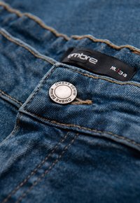 Blaue Denim-Jeans mit einem silbernen Knopf, auf dem der Schriftzug "OMBRE DENIM" steht. Das Etikett trägt die Aufschrift "ombre" und die Größe "34". Sichtbare Nähte.