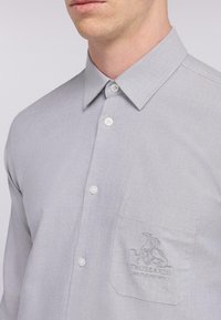 Camisa abotonada de color gris claro hecha de una tela texturizada, que presenta un bolsillo en el pecho con un logo bordado. Cuello clásico y botones blancos.