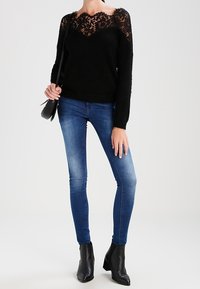Schwarzer flauschiger Pullover mit Spitzenpasse, kombiniert mit blauen Skinny-Jeans und schwarzen Knöchelstiefeln. Das Outfit wird durch eine Umhängetasche ergänzt.