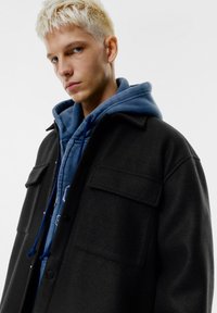 Černá vlněná bunda se dvěma kapsami na prsou, překrytá modrou mikinou s kapucí. Model má krátké, světle blond vlasy.