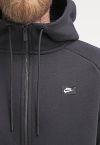 Sudadera con cremallera de Nike en gris oscuro, con un alto cuello, tejido texturizado y un parche de logo negro en el pecho con una cremallera plateada.