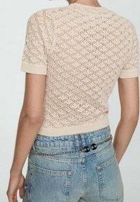 Gebreide crèmekleurige trui met korte mouwen en een diamanten ruitpatroon, gecombineerd met vervaagde blauwe high-waisted jeans en een kettingriem.
