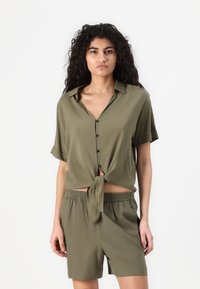 Olive green, short-sleeve shirt with a tied front knot and button closure. Paired with matching loose shorts featuring an elastic waistband.