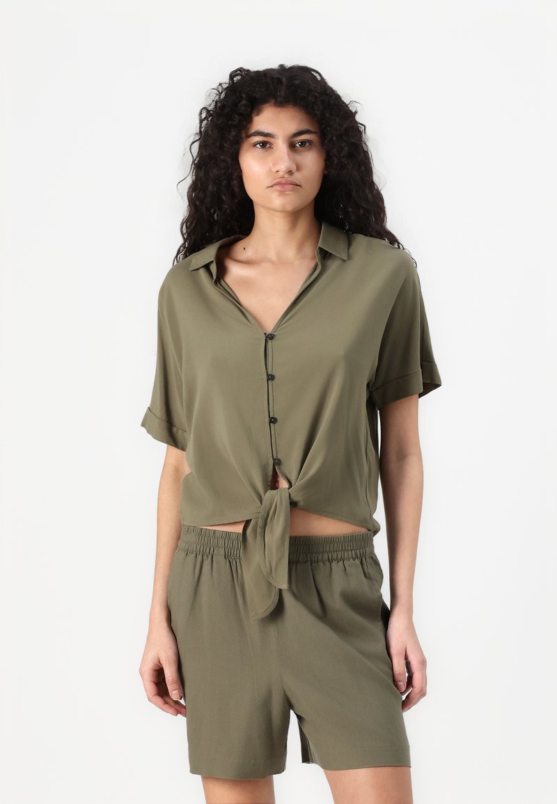 Olive green, short-sleeve shirt with a tied front knot and button closure. Paired with matching loose shorts featuring an elastic waistband.