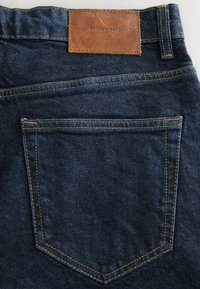 Blugi din denim închise la culoare, cu o etichetă din piele maro pe care scrie "DENIM SUPPLY". Detalii de cusătură pronunțate evidențiază designul buzunarului din spate.