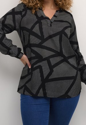 Kvinde med krøllet hår iført en sort-hvid bluse med geometrisk mønster og blå jeans, poserer med den ene hånd på hoften.