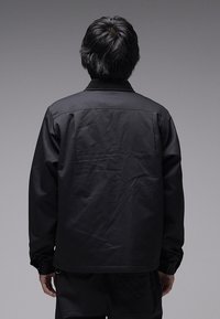 Μαύρο μπουφάν με απλό σχέδιο, που διαθέτει γιακά, μακριά μανίκια και λεία υφή. Η πλάτη είναι ελαφρώς ζαρωμένη, χωρίς ορατό λογότυπο.