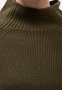 Gros plan d'une personne portant un pull en maille texturé vert olive avec un col montant.