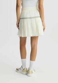 Helles cremefarbenes, plissiertes Kleid mit strukturiertem Taillenbund, der einen schwarzen Streifen aufweist. Kombiniert mit sneakers mit goldenen Akzenten und weißen Socken.