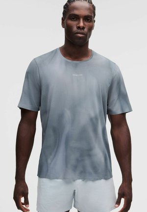 Hombre con el pelo trenzado lleva una camiseta deportiva gris clara y pantalones cortos blancos, de pie frente a un fondo blanco liso, mirando hacia adelante.