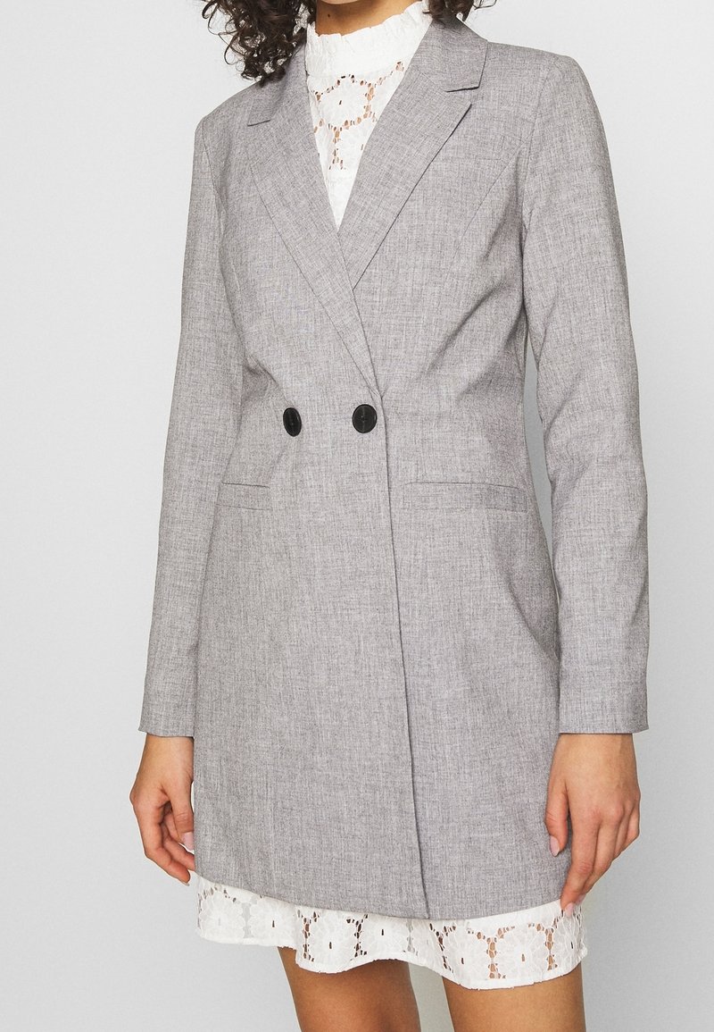 Szary, teksturowany żakiet z dwoma czarnymi guzikami, w dopasowanym kroju, z długimi rękawami, noszony na białej koronkowej sukience z falbanii u dołu.