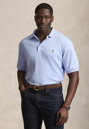 Mand iført en lyseblå poloshirt, mørke jeans, brunt bælte og armbåndsur, står med hænderne i lommerne mod en neutral baggrund.