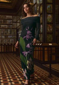 Mørkegrøn off-the-shoulder kjole med stort lilla blomsterprint, tætsiddende overdel og svajende nederdel. Glat tekstur, parret med sorte sandaler.