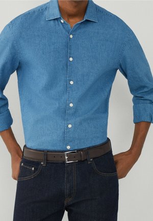 Mann trägt ein hellblaues Hemd mit langen Ärmeln und aufgerollten Ärmeln, dunkelblaue Jeans und einen braunen Ledergürtel.
