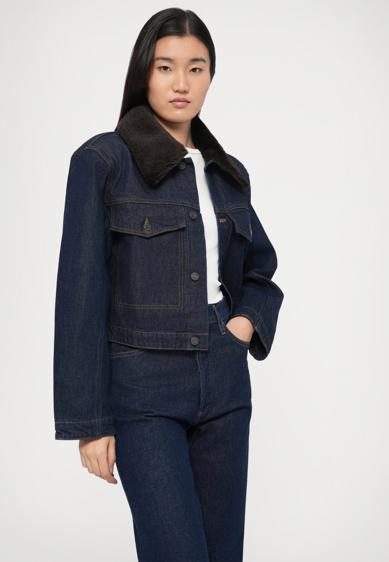 Dunkle Jeansjacke mit braunem Pelzkragen, versehen mit Knopfverschluss, zwei Brusttaschen und einem kurzen Schnitt.