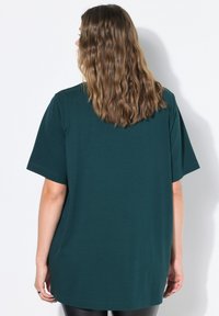 Žena s vlnitými světle hnědými vlasy se otáčí, na sobě má tmavě zelené oversized tričko s krátkým rukávem na bílém pozadí.