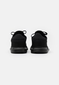 Des chaussures de sport noires avec une tige en mesh texturé, un talon arrondi et des languettes à l'arrière. La semelle est plate et lisse.