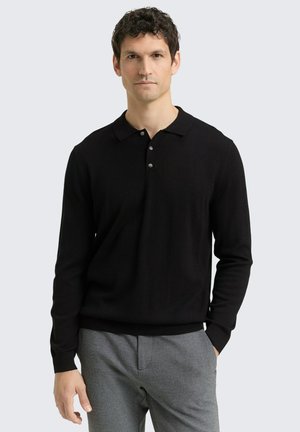 Mann mit dunklen lockigen Haaren, der ein schwarzes langärmeliges Poloshirt und graue Hose trägt, steht mit einer Hand in der Tasche vor einem einfarbigen Hintergrund.