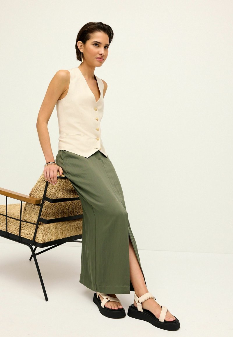 Débardeur beige sans manches avec des boutons dorés, associé à une longue jupe verte avec une fente sur le côté. Sandales à plateforme noires avec des brides beiges.