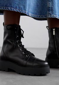 Bottines en cuir noir avec bouts ronds. Présentent des œillets blancs proéminents, des lacets, des semelles noires épaisses et un détail de fermeture éclair sur le côté.