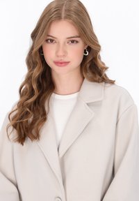 Cappotto oversize grigio chiaro con revers e chiusura a un bottone. Tessuto morbido. Indossato sopra un top bianco a coste.