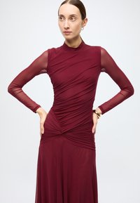 Burgundy dress made of textured fabric, featuring long sheer sleeves, draped design, and a knotted waist detail for a sleek silhouette.