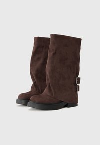 Braune Wildleder-Stiefel bis zur Wade mit einem lässigen Design, schwarzen Gummisohlen und metallicfarbene Schnallen an der Seite. Glatte Oberfläche.