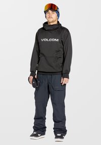 Black hooded sweatshirt with "VOLCOM" logo, paired with dark blue snow pants. Model holds black gloves, wearing ski goggles and sneakers.