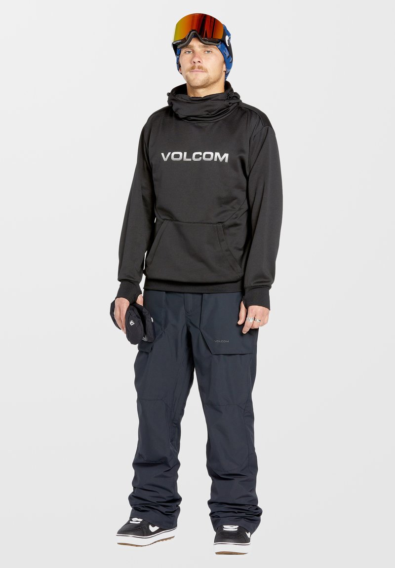 Black hooded sweatshirt with "VOLCOM" logo, paired with dark blue snow pants. Model holds black gloves, wearing ski goggles and sneakers.