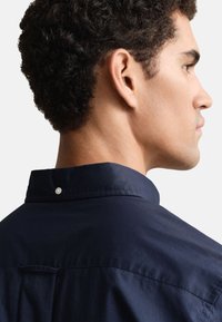 Camisa de algodão azul-marinho com colarinho abotoado e um pequeno detalhe de botão na nuca, textura suave e corte ajustado.