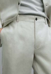 Pantaloni in lino beige con fronte piatto, chiusura a un bottone e tasche laterali diagonali. Texture liscia con una leggera lucentezza.