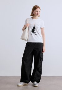 Λευκό γραφικό t-shirt με σχέδιο πόλης, συνδυασμένο με μαύρο cargo παντελόνι με μεγάλες τσέπες και χαλαρή εφαρμογή. Ελαφριά τσάντα και ασημένια αθλητικά παπούτσια.