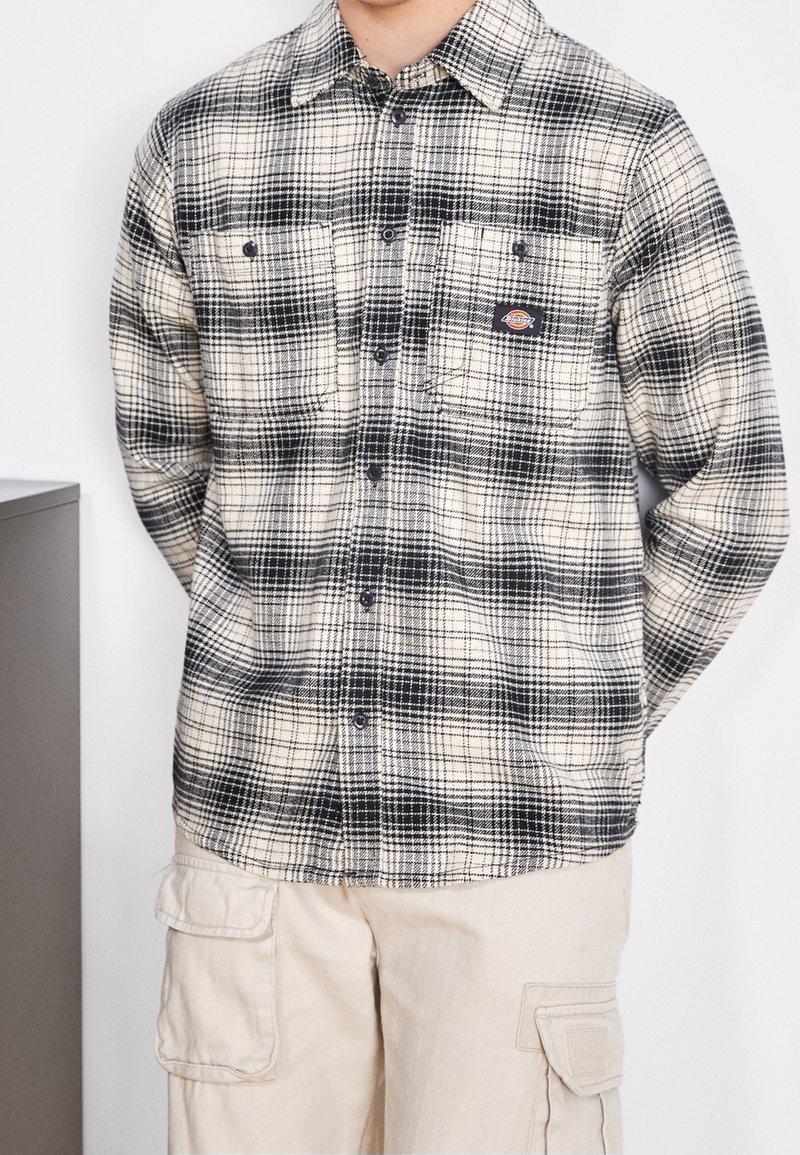 Personne portant une chemise à carreaux noire et blanche boutonnée avec deux poches poitrine et un pantalon cargo beige avec poches latérales.