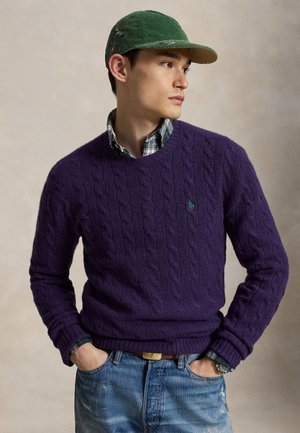 Giovane uomo con un maglione viola a trecce, un berretto di velluto verde, una camicia a quadri e jeans blu sbiaditi, che guarda di lato con le mani nelle tasche.