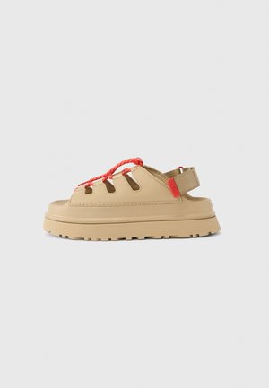 Sandalias beige con suela de goma texturizada, con secciones recortadas, cordones trenzados rojos y una correa trasera para asegurar.