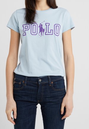Lichtblauw katoenen T-shirt met "POLO" in paars bedrukt, met een motief van een ruiter te paard. Draagt donkerblauwe spijkerbroek. Korte mouwen, ronde hals.