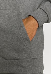 Main glissée dans la poche avant d'un sweat-shirt gris à manches longues avec poignets côtelés et tissu texturé.