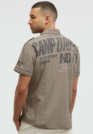 Young man with short hair wearing a gray short-sleeve shirt with bold "CAMP DAVID" text and graphics on the back, hands in pockets.