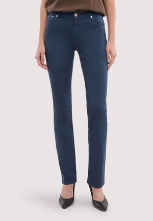 Frau trägt dunkelblaue Slim-Fit-Hose und schwarze High-Heels, steht vor einem einfarbigen hellen Hintergrund.