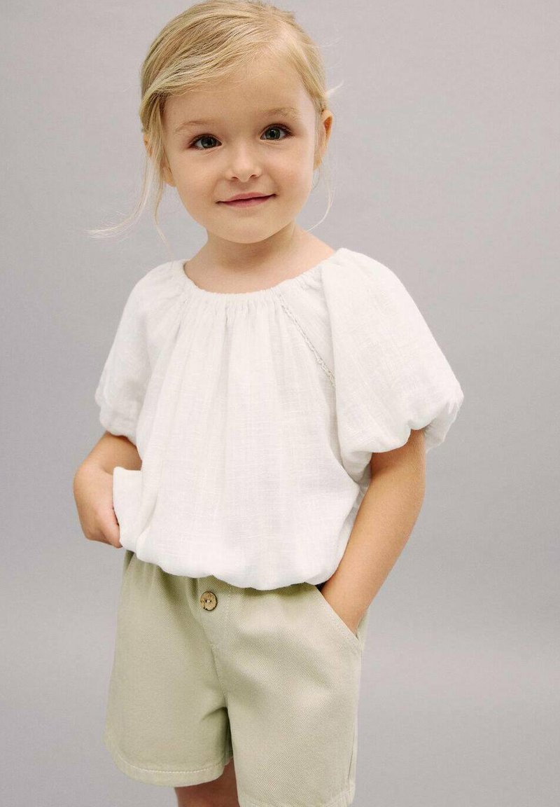 Blusa bianca a maniche corte con scollatura arricciata, abbinata a pantaloni corti beige chiaro con chiusura a bottone e tasche laterali.