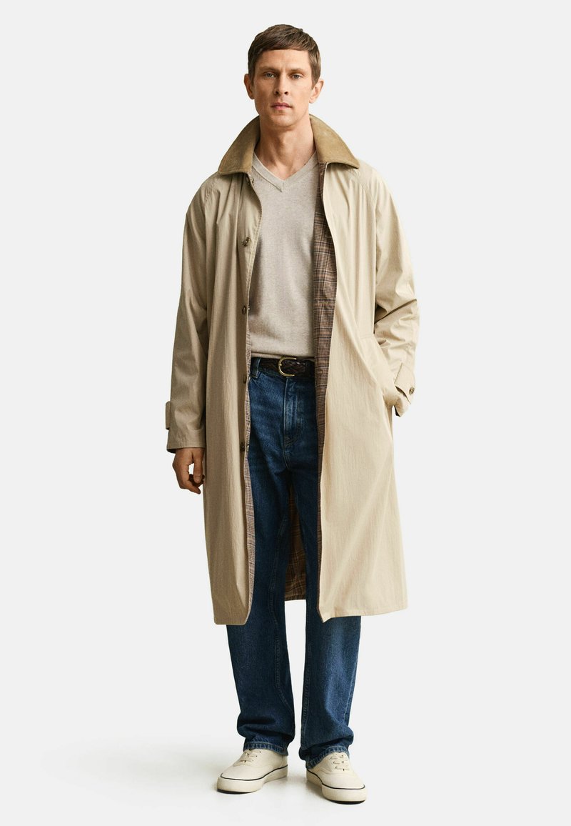 Mann steht und trägt einen beigen langen Mantel über einem hellgrauen V-Ausschnitt-Pullover, blaue Jeans, schwarzen Gürtel und weiße Turnschuhe vor weißem Hintergrund.